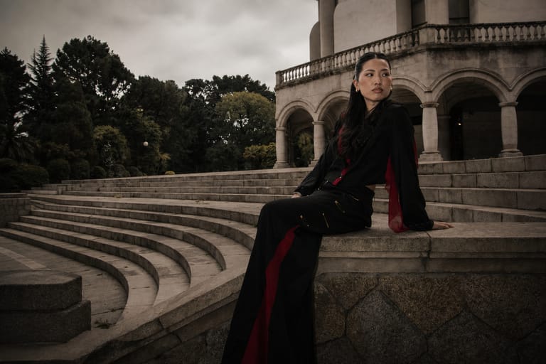A model seated on curved stone steps in front of a neoclassical arcade, wearing a black II Acto look with a red lining and red side stripe.