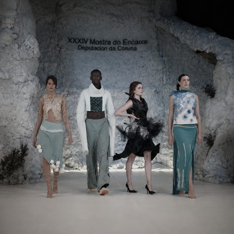 Four Acqua Vitae looks on the runway of the XXXIV Mostra do Encaixe de Camariñas, against a white rock backdrop: knotted mesh, sea-green tailoring, a black feathered dress and a floral-embroidered top.